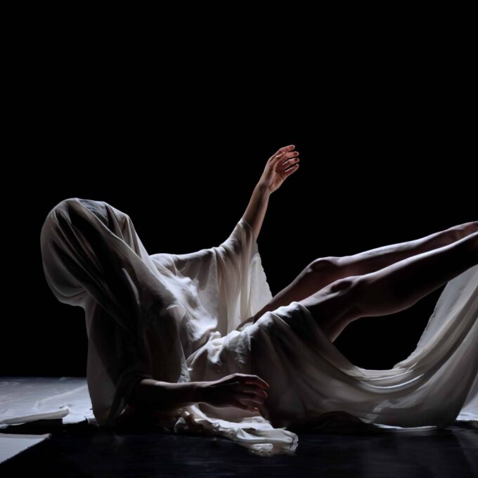 Sur un fond noir, un corps est allongé au sol dans une pose contorsionnée. Il est entièrement drapé d'un tissu blanc et vaporeux qui cache le visage, ne laissant apparaître qu'un bras et les jambes. L'éclairage est dramatique et met en valeur les formes du corps et les plis du tissu.
