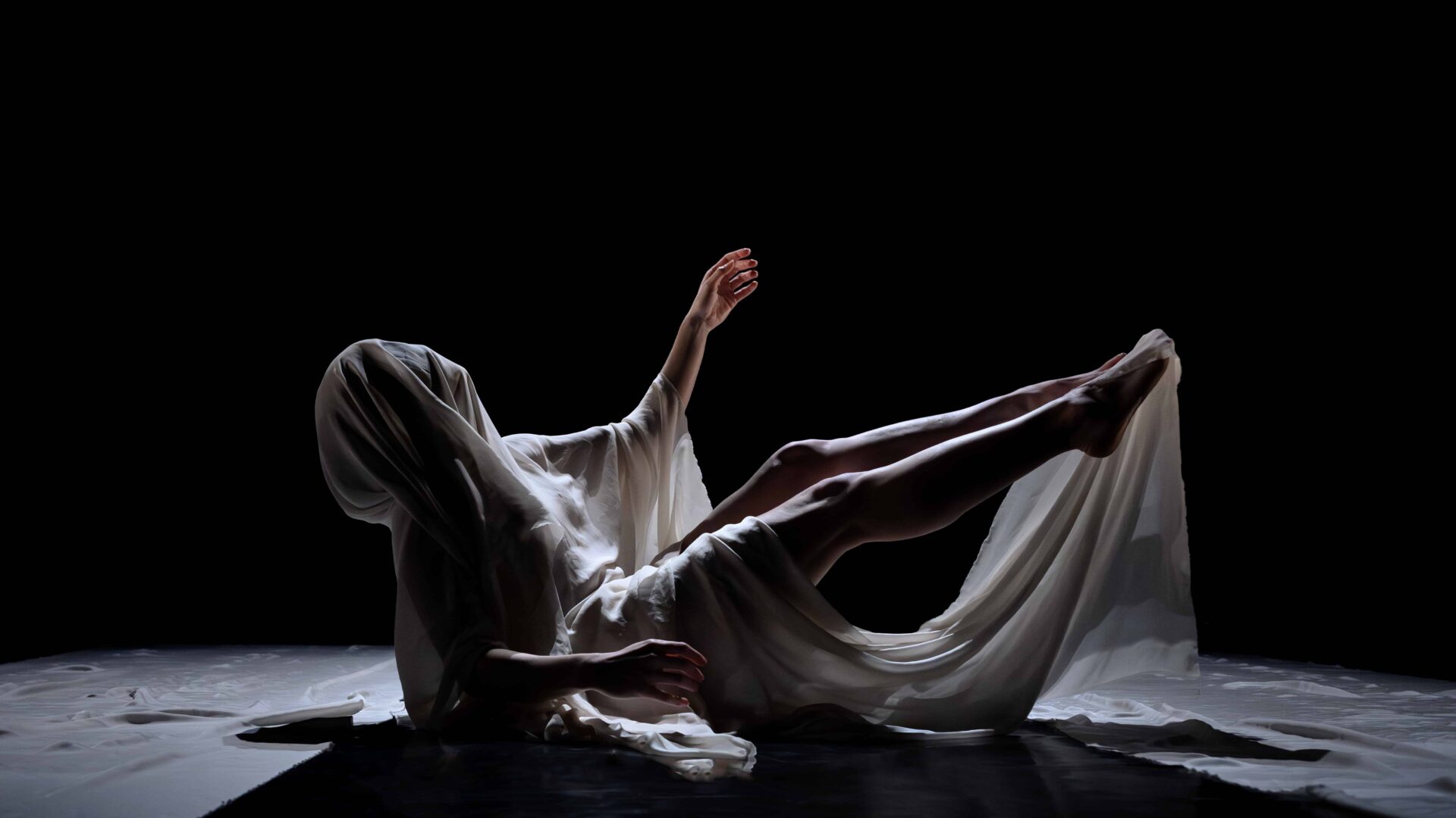 Sur un fond noir, un corps est allongé au sol dans une pose contorsionnée. Il est entièrement drapé d'un tissu blanc et vaporeux qui cache le visage, ne laissant apparaître qu'un bras et les jambes. L'éclairage est dramatique et met en valeur les formes du corps et les plis du tissu.